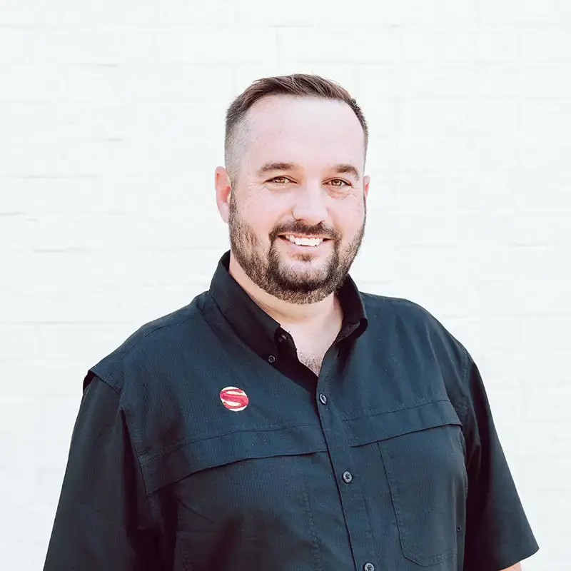 A smiling man with short hair and a beard, wearing a black button-up shirt with a red circular pin, stands in front of a plain white brick wall.