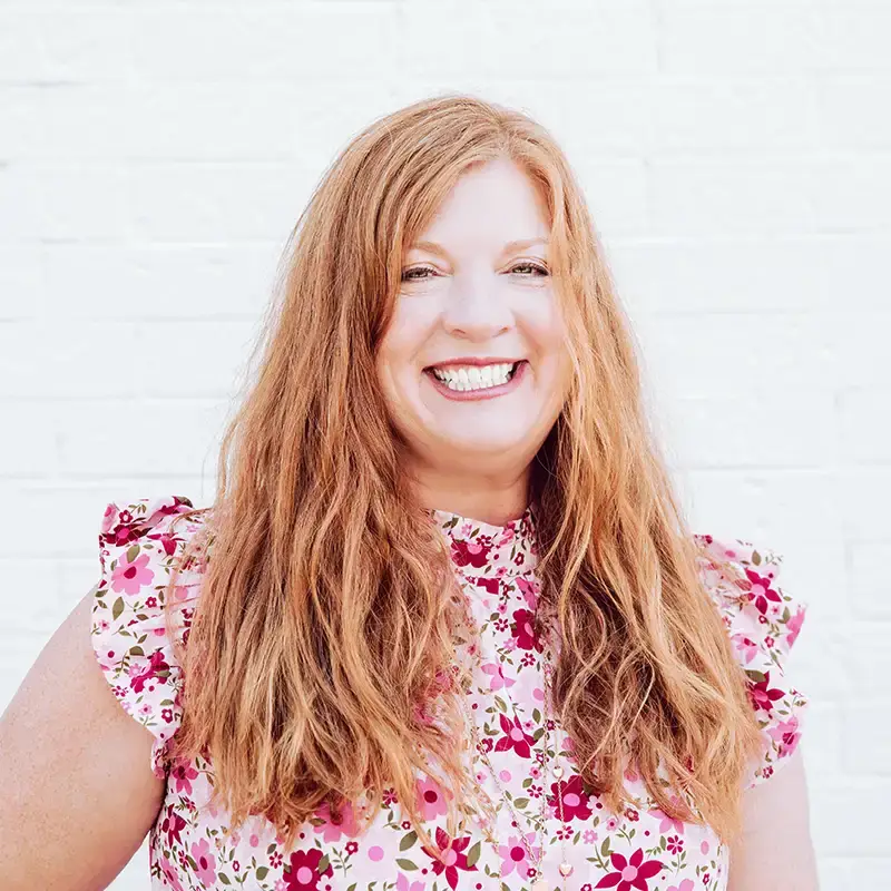 A woman with long, wavy red hair smiles widely while wearing a pink floral blouse with ruffled sleeves, standing in front of a white brick wall.