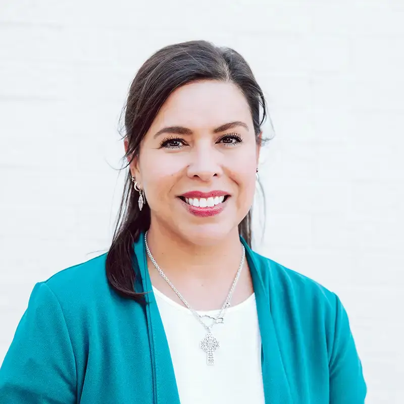 A woman with long dark hair wearing a teal blazer, white top, silver earrings, and a necklace, smiling in front of a light-colored background.