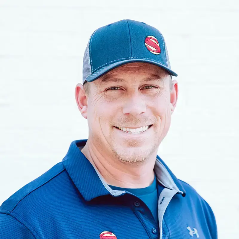 A man wearing a blue baseball cap and a blue collared shirt with red and white logos smiles at the camera against a plain, light background.