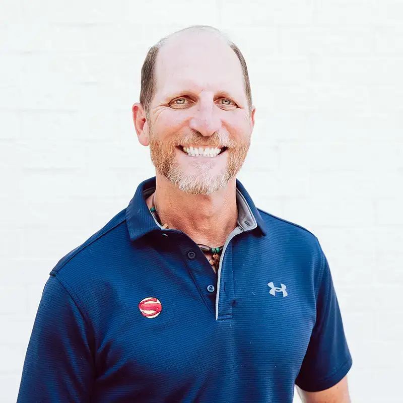 A smiling man with short hair and a beard wearing a navy blue Under Armour polo shirt and a red pin, standing in front of a plain white background.
