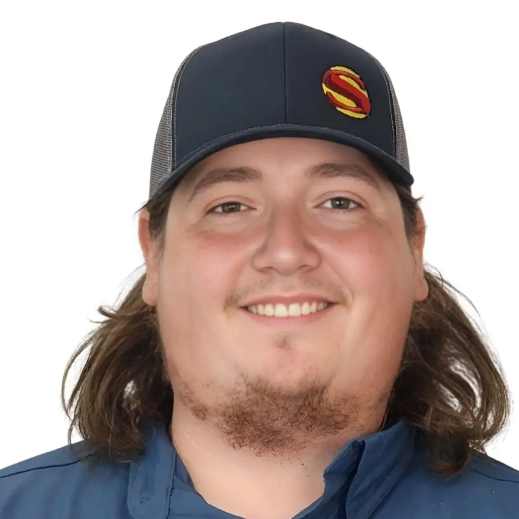 A person with long brown hair, wearing a blue cap with a red and yellow S logo, and a blue shirt, is smiling at the camera against a plain white background.