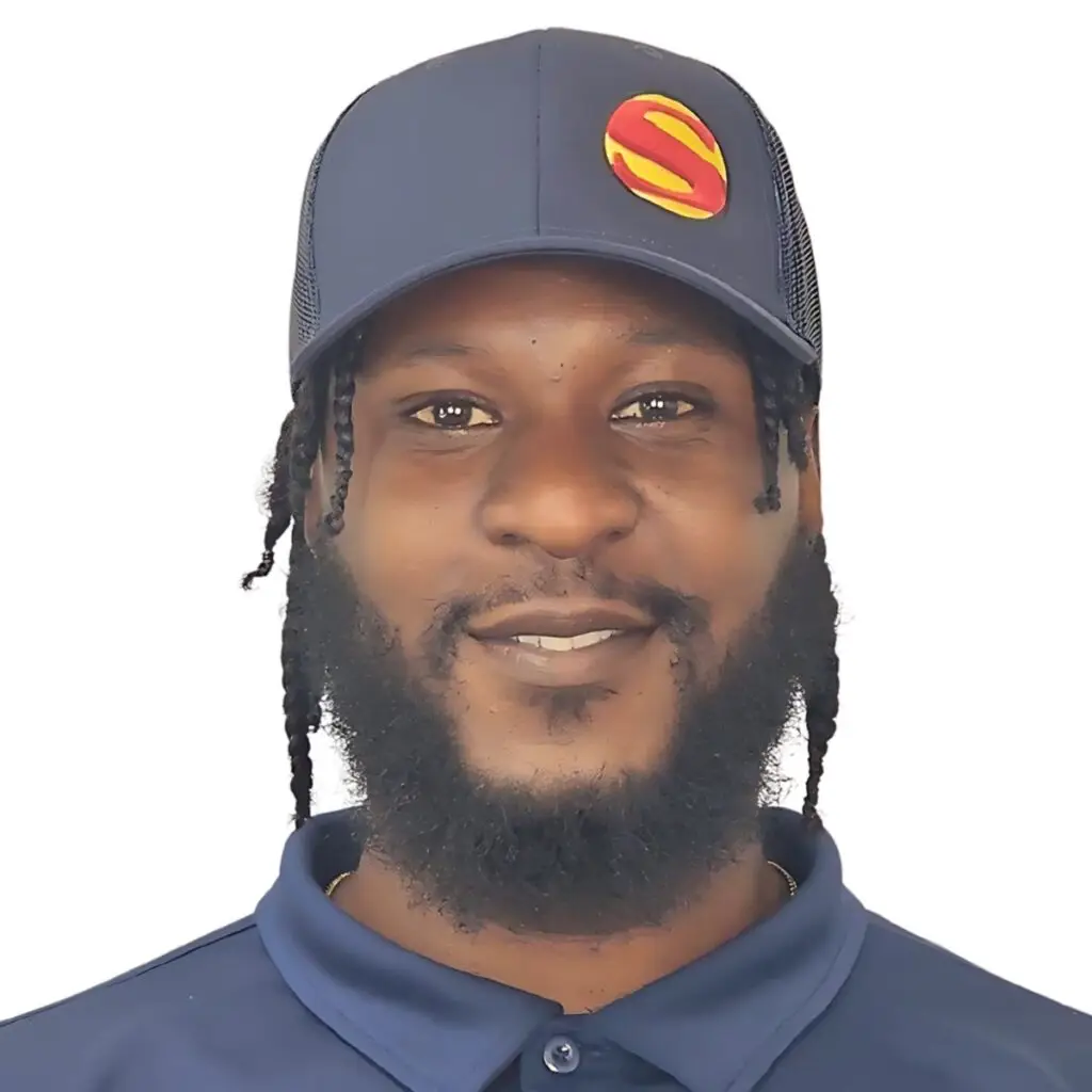 A man with a beard and braided hair smiles, wearing a navy blue cap with a red and yellow S logo and a matching collared shirt, set against a plain white background.