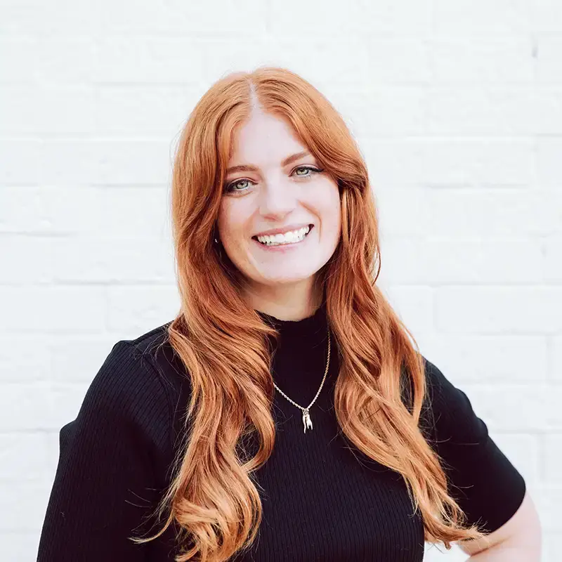A woman with long red hair and a black top smiles in front of a white brick wall. She wears a gold necklace with a small pendant.